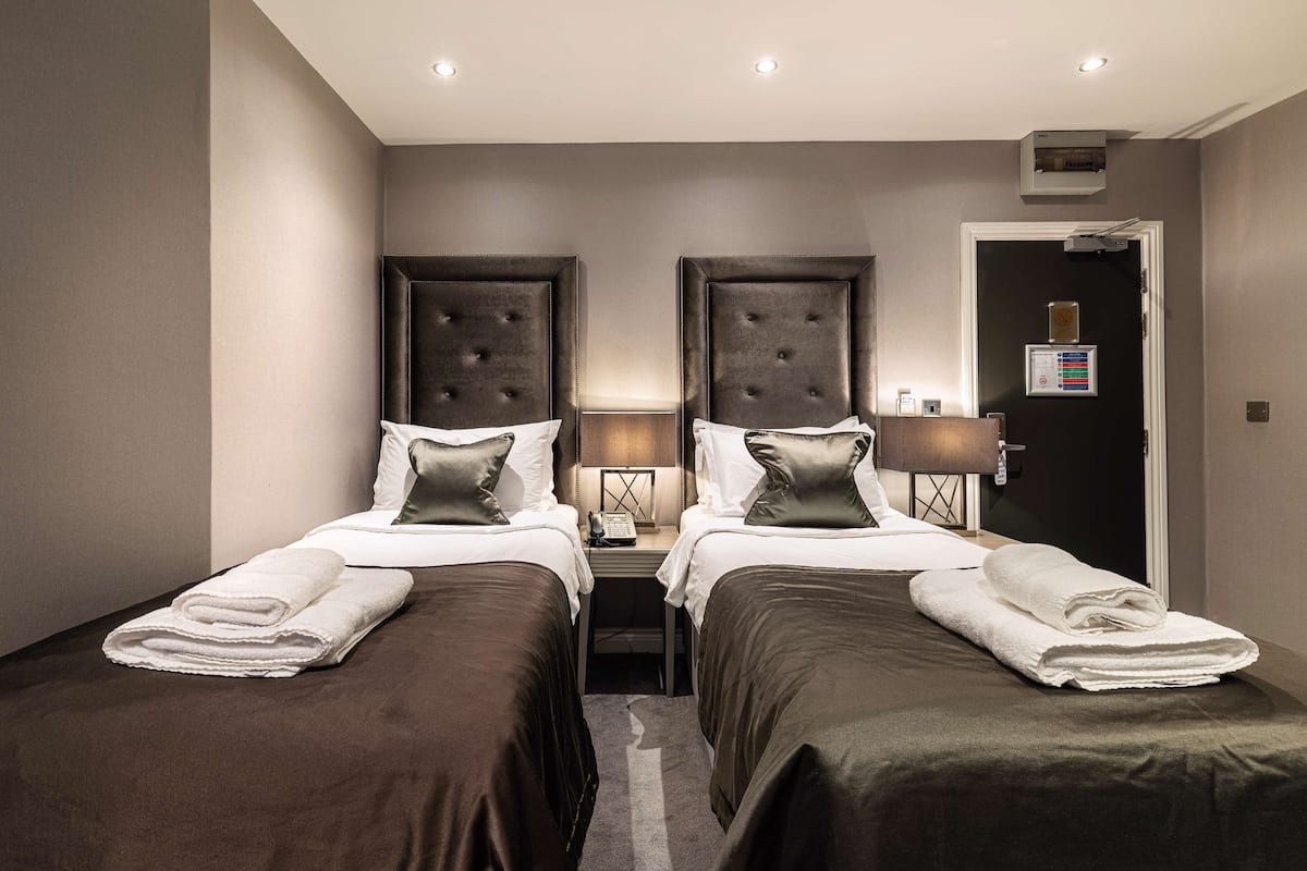 A hotel room with two beds, each with a headboard, bedside tables with lamps, and a door with a control panel.