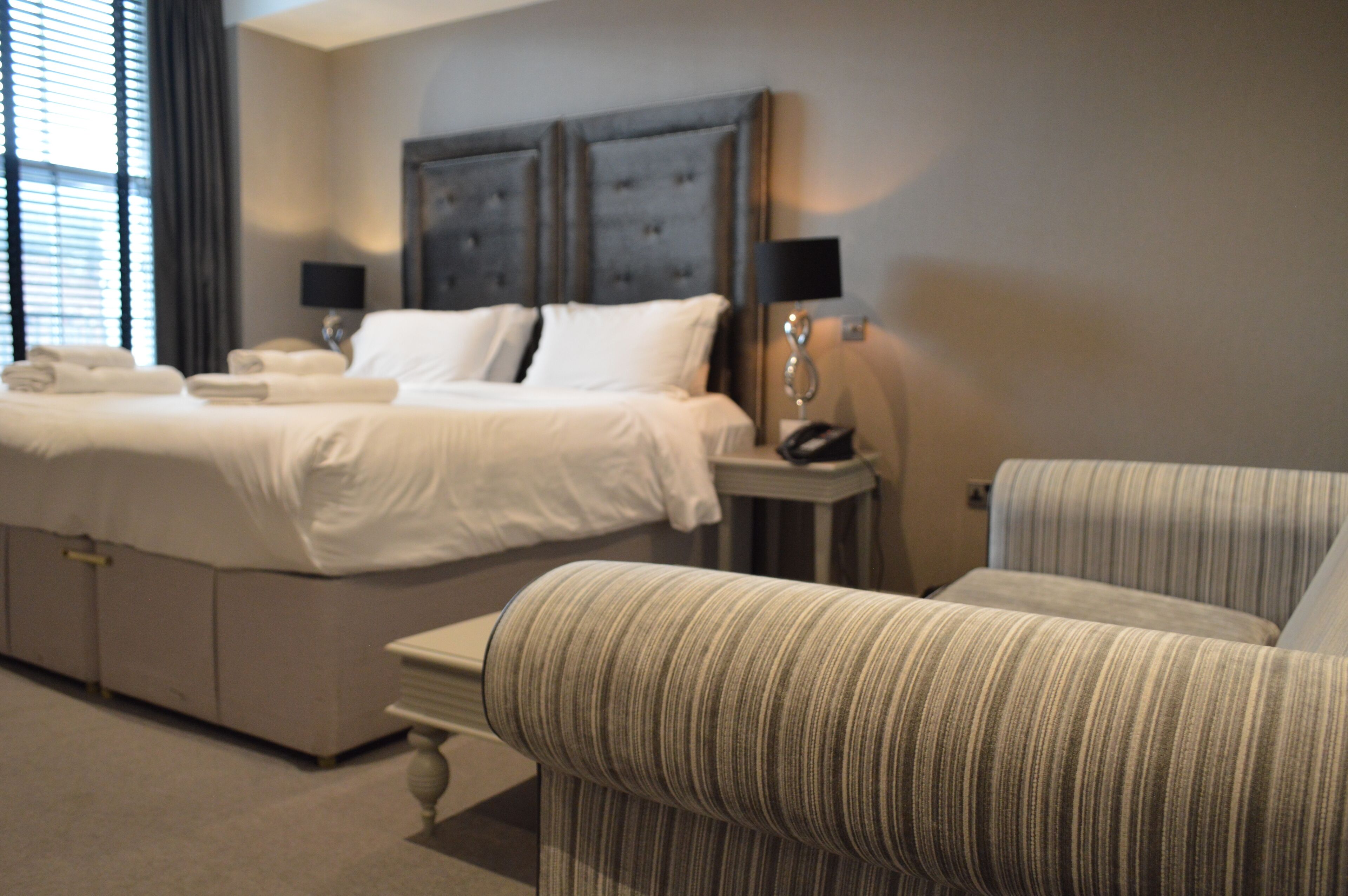 A hotel room with a large bed, a bedside table, a lamp, and a striped armchair.