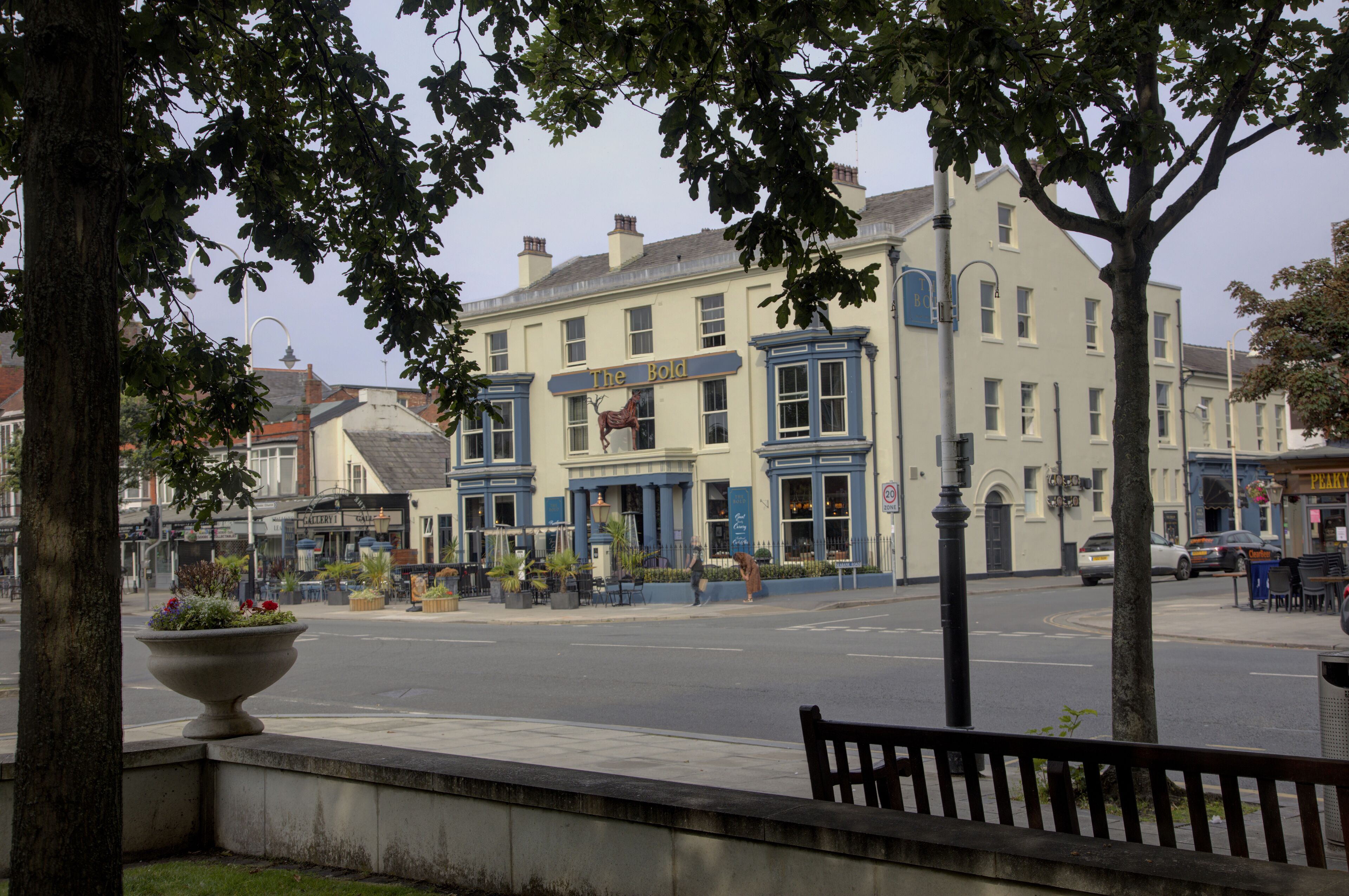 A street view with a building labeled "The Bold" featuring a traditional facade, a large planter with flowers, and a bench along the sidewalk.