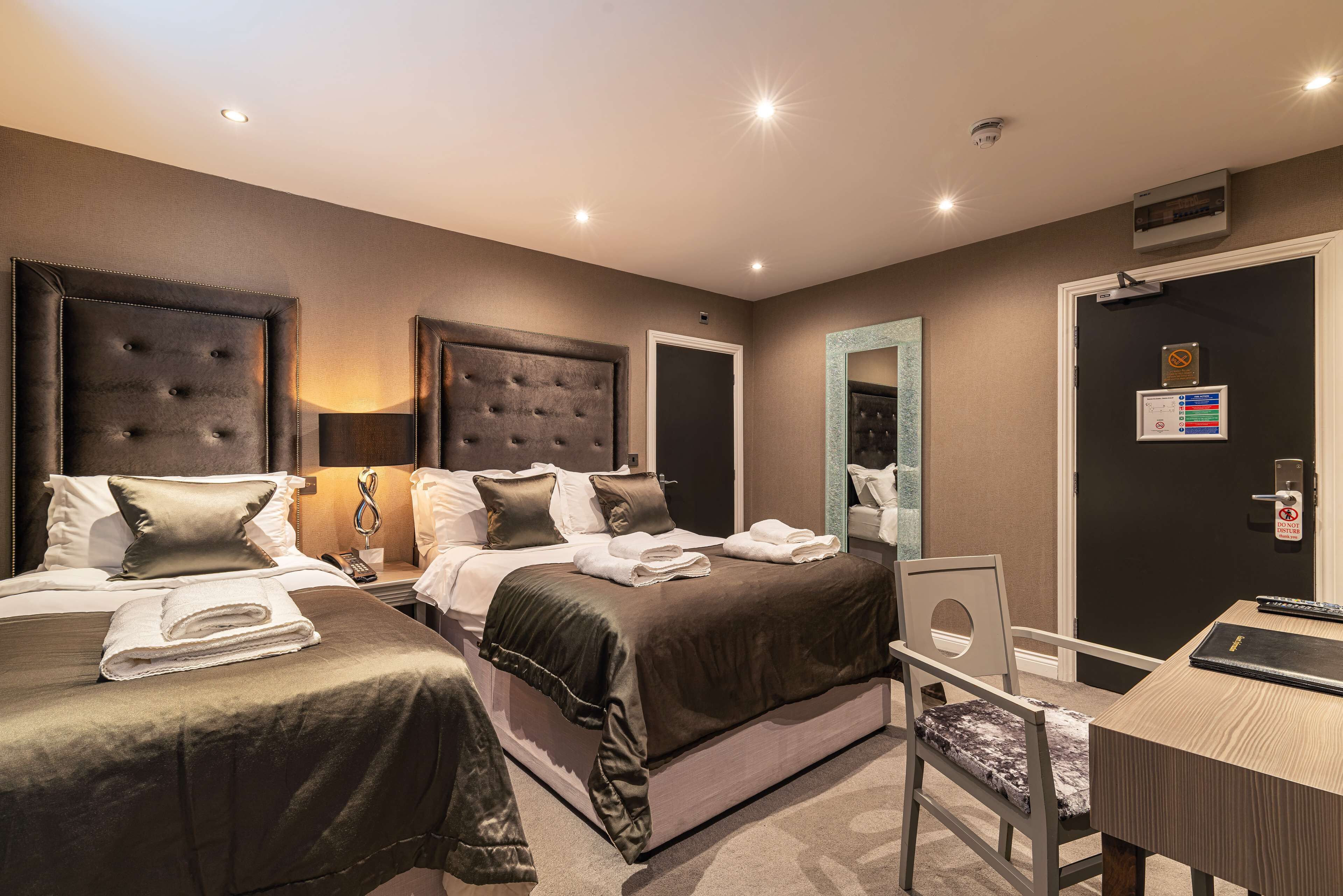 A hotel room with two beds, a desk, a chair, and a door with a security camera.