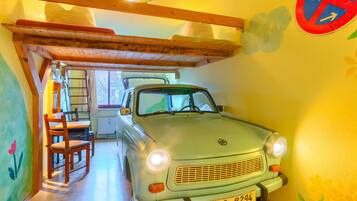 A vintage car parked inside a room with wooden beams on the ceiling, a dining table with chairs, and a window with a view of trees.