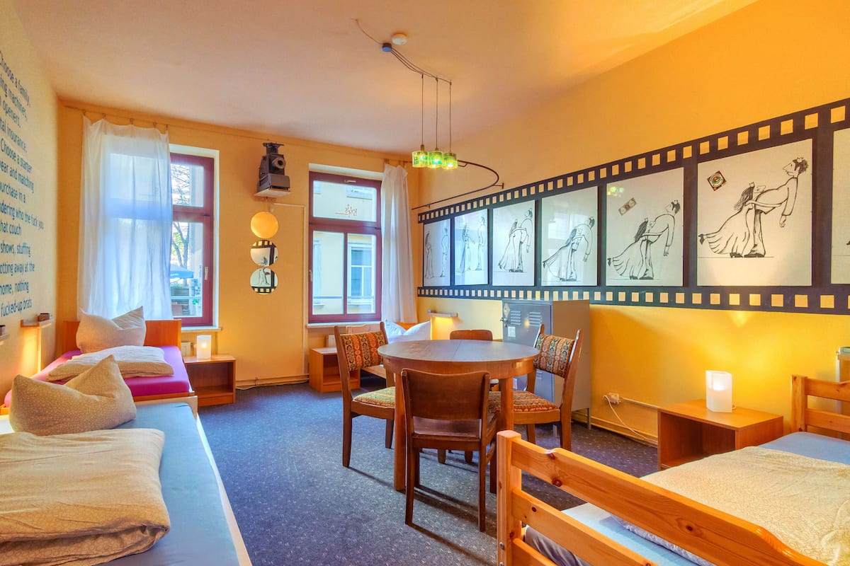 A cozy hotel room with two beds, a dining table, and chairs. There are curtains on the windows, and artwork on the wall.