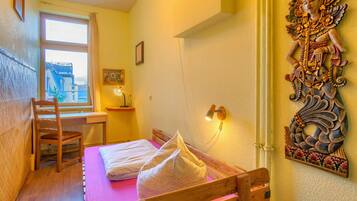 Un dormitorio con una cama de madera, un escritorio y una silla. Se ve una ventana con cortinas, una lámpara de pared y un adorno en la pared.
