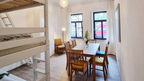 A room with a bunk bed, a dining table with chairs, a potted plant, and two windows with curtains. - Lollis Homestay - Hostel (Dresden)