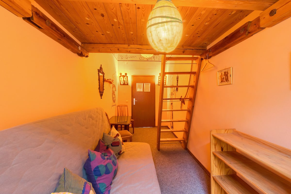 A cozy room with a wooden ceiling, a sofa with colorful pillows, a wooden ladder, and a small table with chairs.