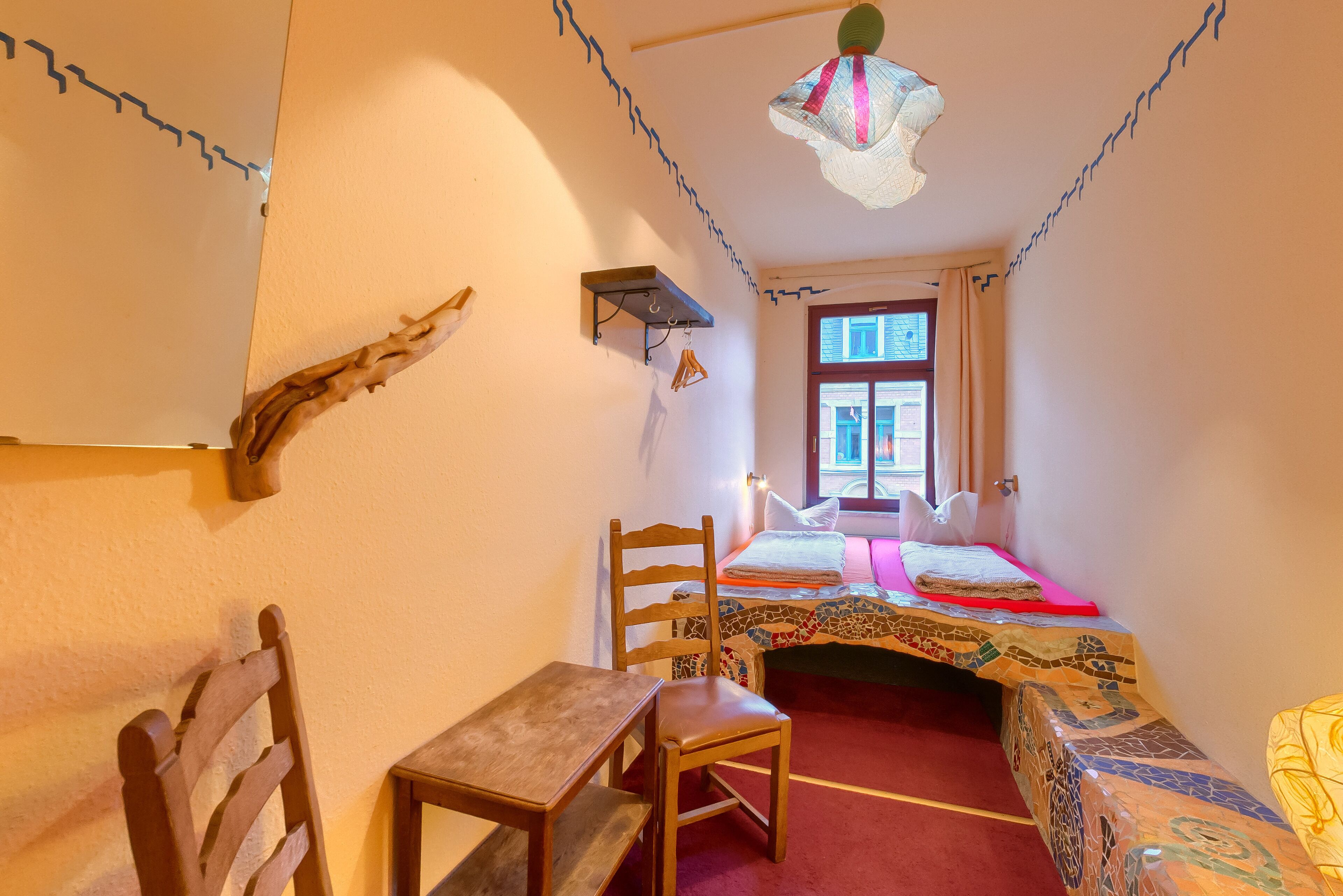 Una habitación con una cama, una silla de madera, una mesita, un estante y una ventana.