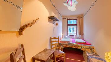 Una habitación con una cama, una silla de madera, una mesita, un estante y una ventana.