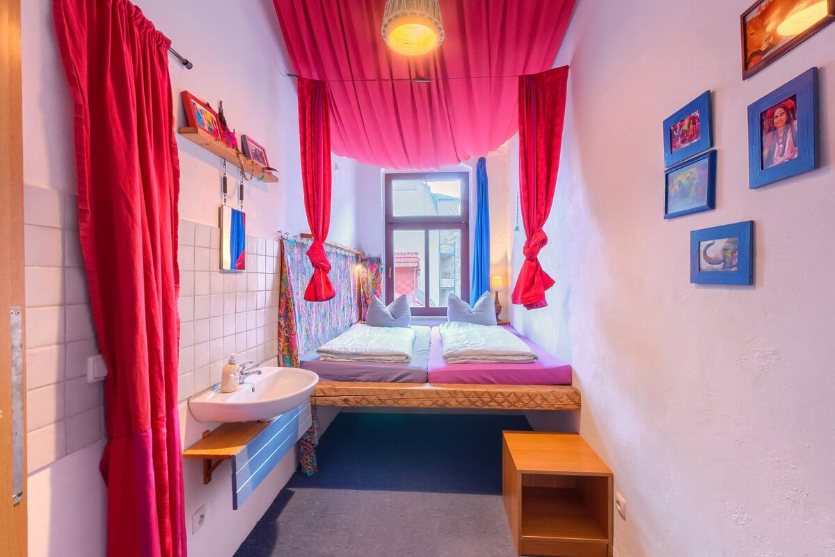 A small, brightly lit room with a bed, a sink, and a window.