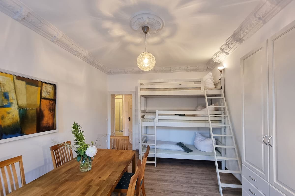 A room with a bunk bed, a wooden dining table with chairs, a vase with flowers, and a painting on the wall.