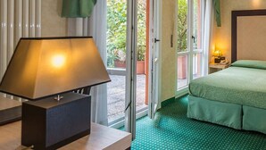 A hotel room with a bed, a desk with a lamp, and a view of greenery through the door. - Don Abbondio (Lecco)