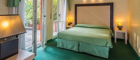 Habitación de hotel con cama, escritorio con lámpara y vista a la vegetación por la puerta.