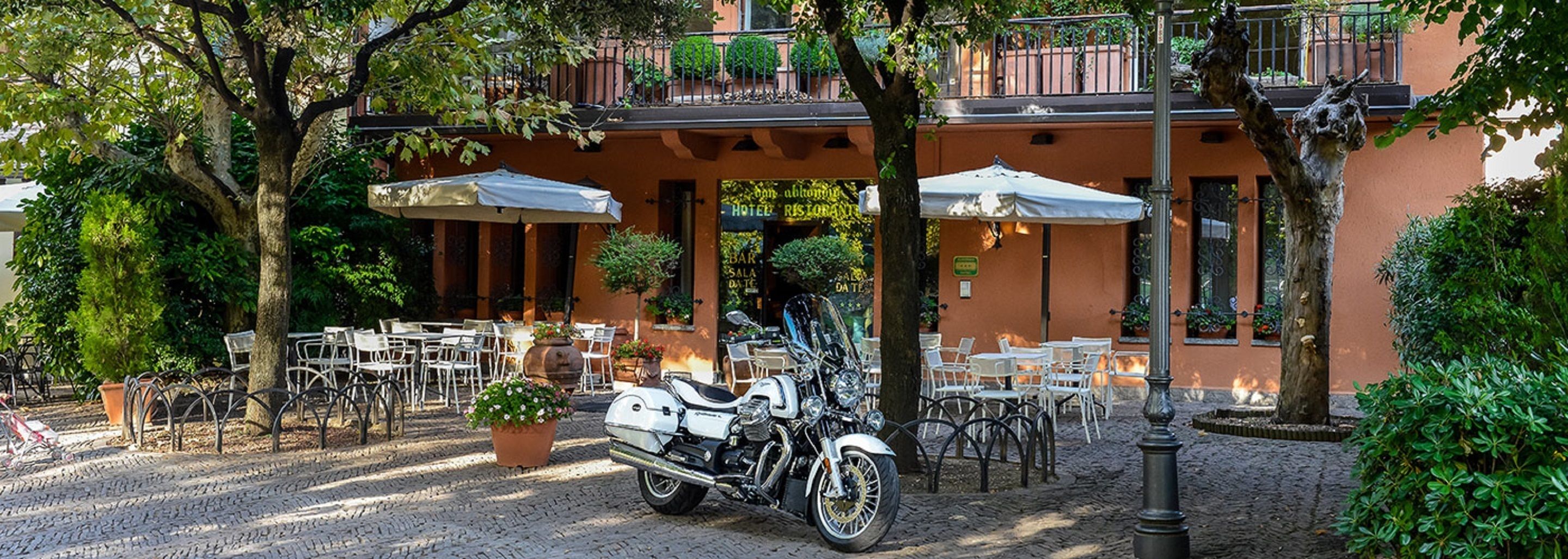 En brostensbelagt terrasse med udendørs møbler, en parkeret motorcykel og en bygning med altan.