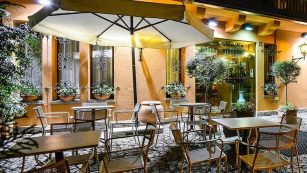 An outdoor cafe area with tables and chairs, potted plants, and a stone floor. - Don Abbondio (Lecco)