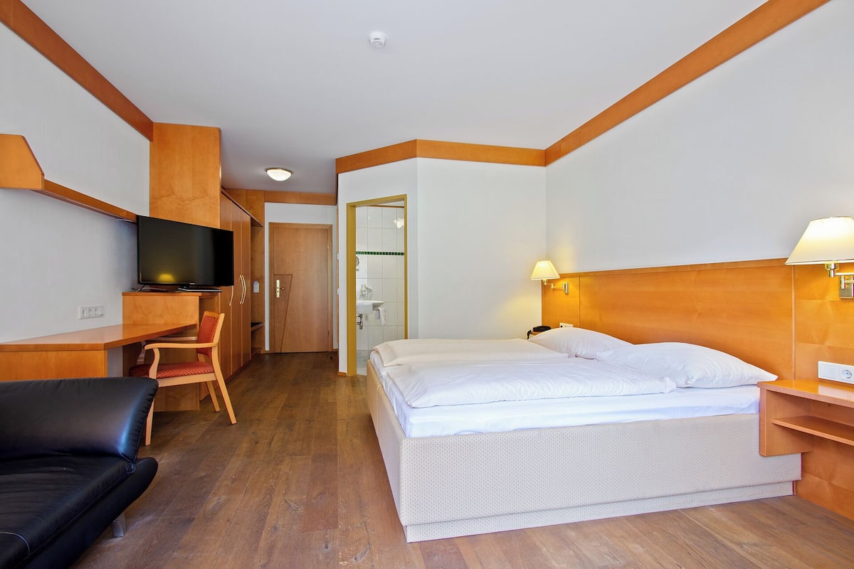 A hotel room with a bed, a desk with a TV, a chair, and a bathroom visible through an open door.
