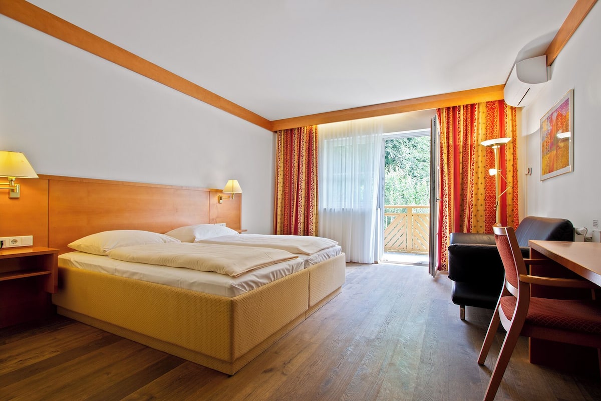 A hotel room with a double bed, a desk, a chair, a lamp, and a view of the outdoors.