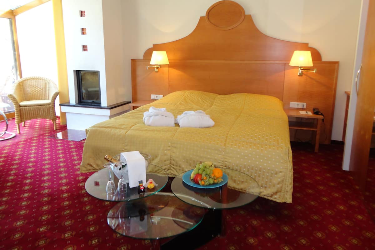 A hotel room with a large bed, two bedside lamps, a small table with a fruit bowl, a glass table with wine glasses, and a wicker chair.