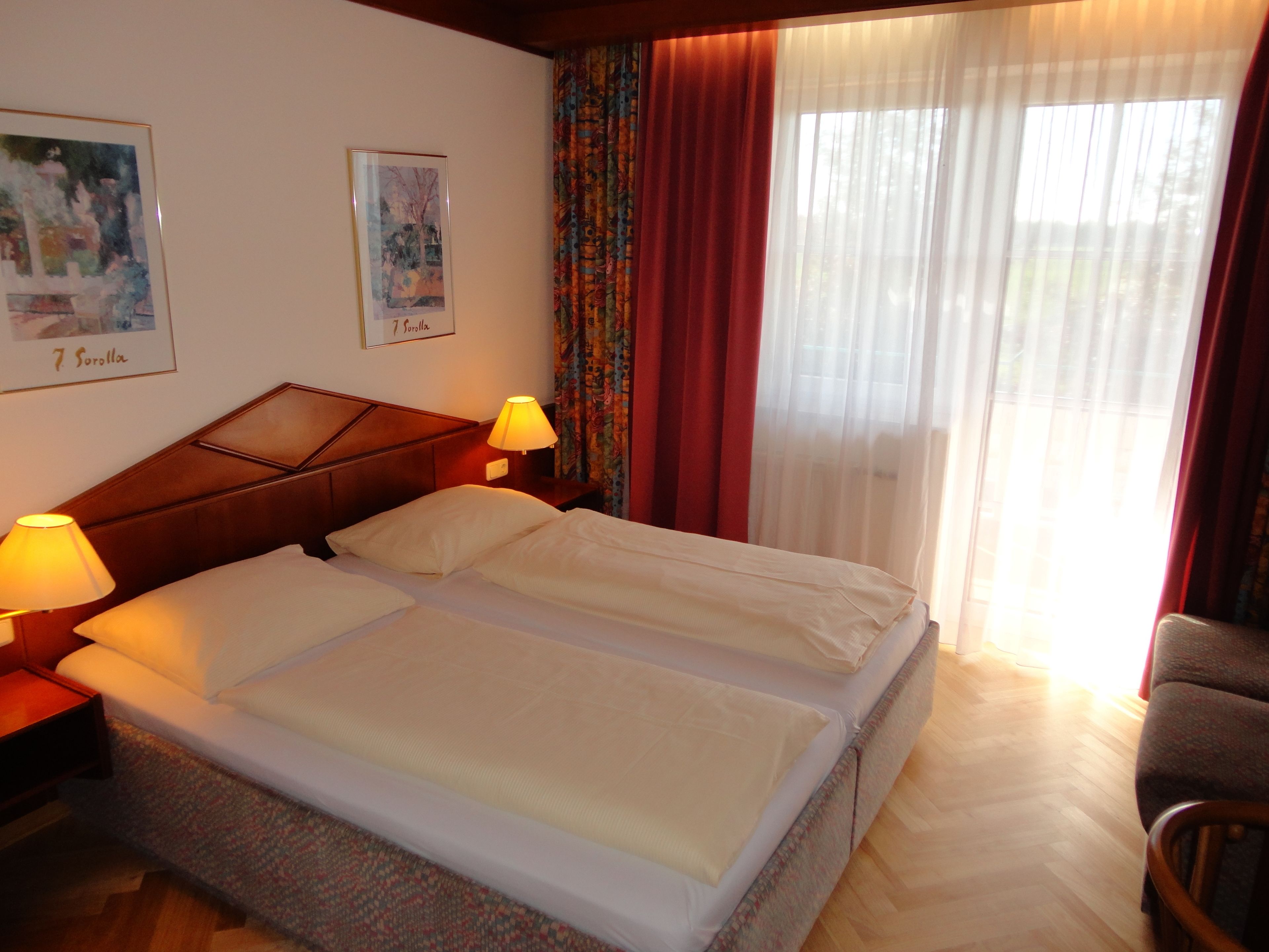 Ein Hotelzimmer mit Doppelbett, zwei Nachttischlampen, einem Holz-Kopfteil, einem Sessel und einem Fenster mit durchsichtigen Vorhängen.
