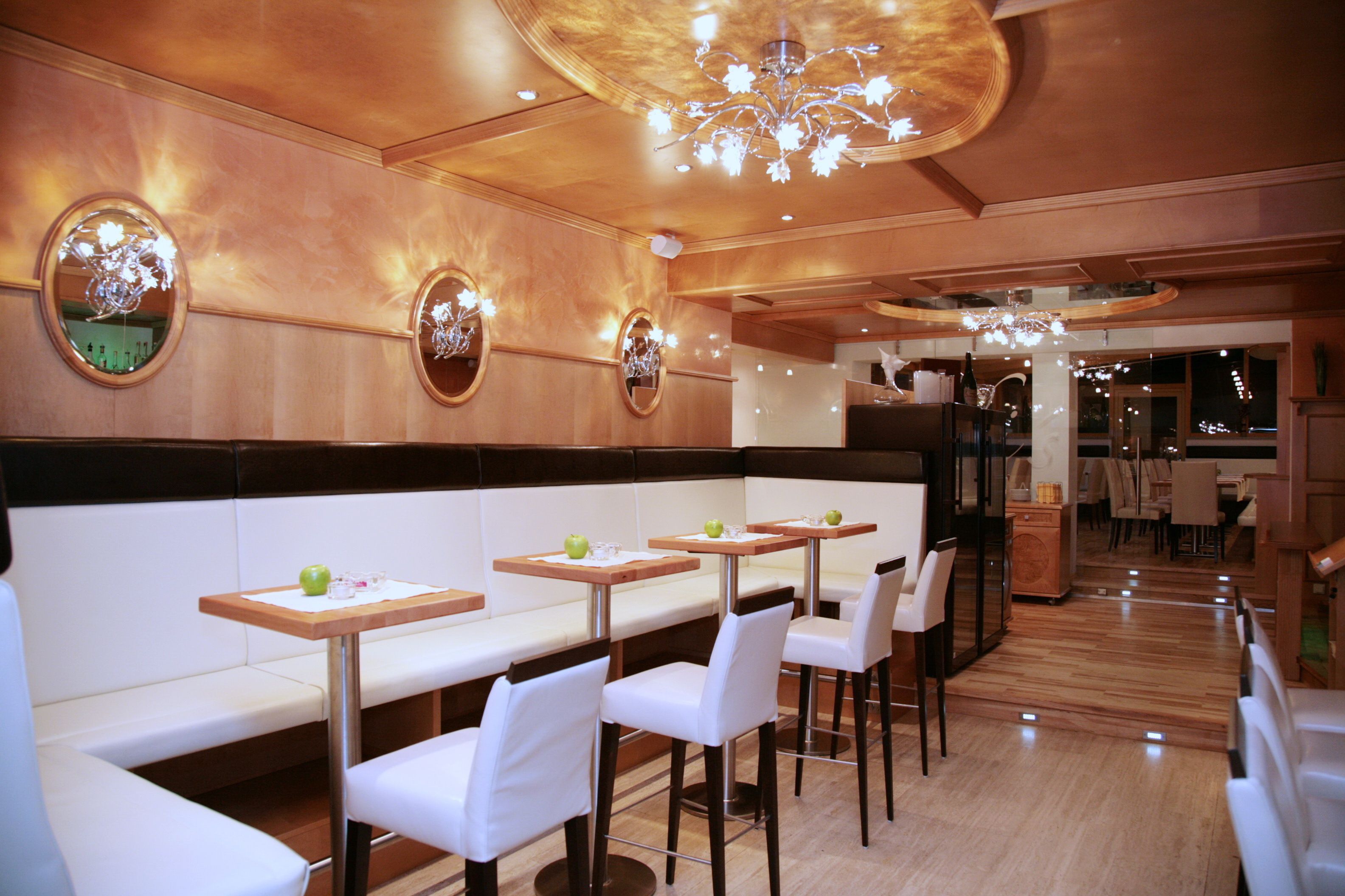 Un restaurante con paredes de paneles de madera, espejos redondos y una lámpara de araña. Las mesas están dispuestas con sillas blancas y manzanas verdes.