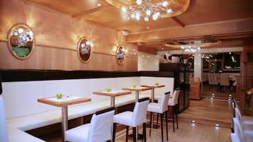 Un restaurante con paredes de paneles de madera, espejos redondos y una lámpara de araña. Las mesas están dispuestas con sillas blancas y manzanas verdes.