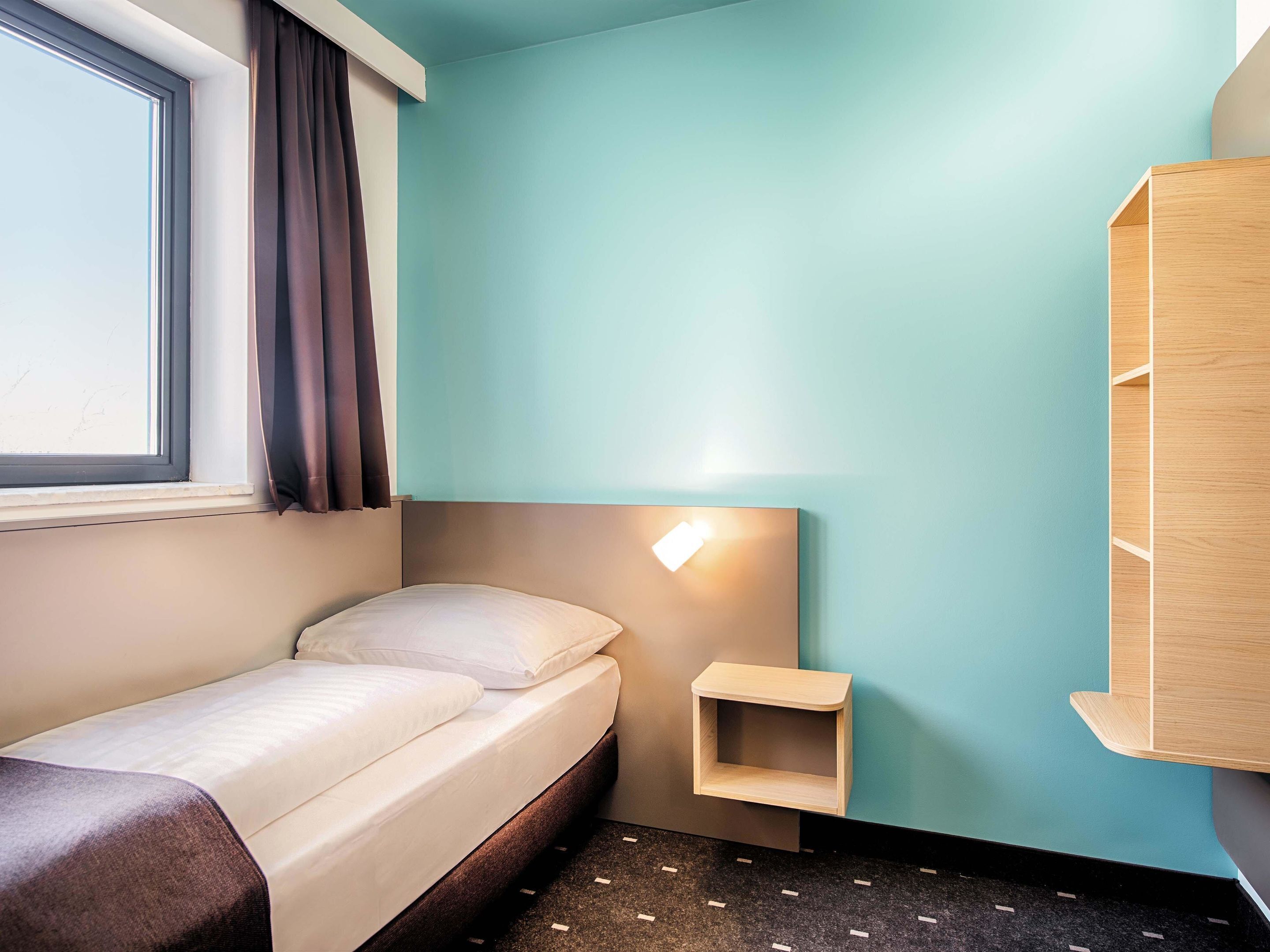 Ein kleines, ordentlich eingerichtetes Schlafzimmer mit einem Einzelbett, einem Nachttisch, einer Wandleuchte und einem Fenster mit Vorhängen.