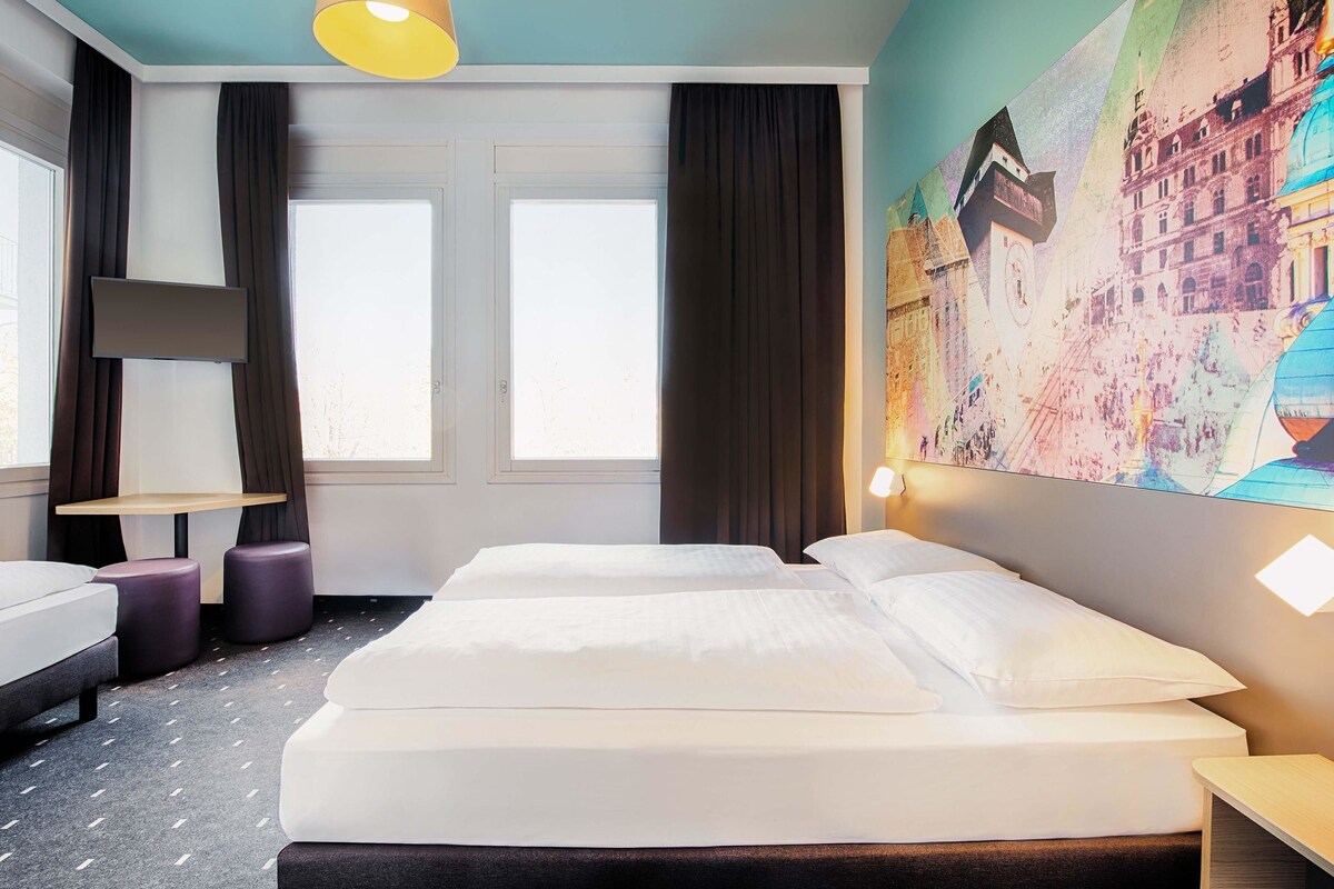 A hotel room with a bed, two bedside tables, a desk, and a large window.