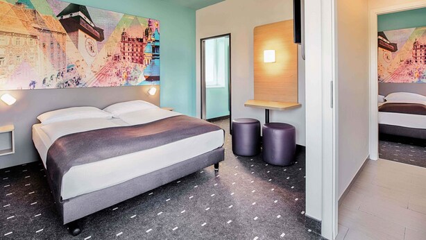 Une chambre d’hôtel avec un lit, une lampe fixée au mur, une œuvre d’art représentant une carte et une porte donnant sur une autre pièce.