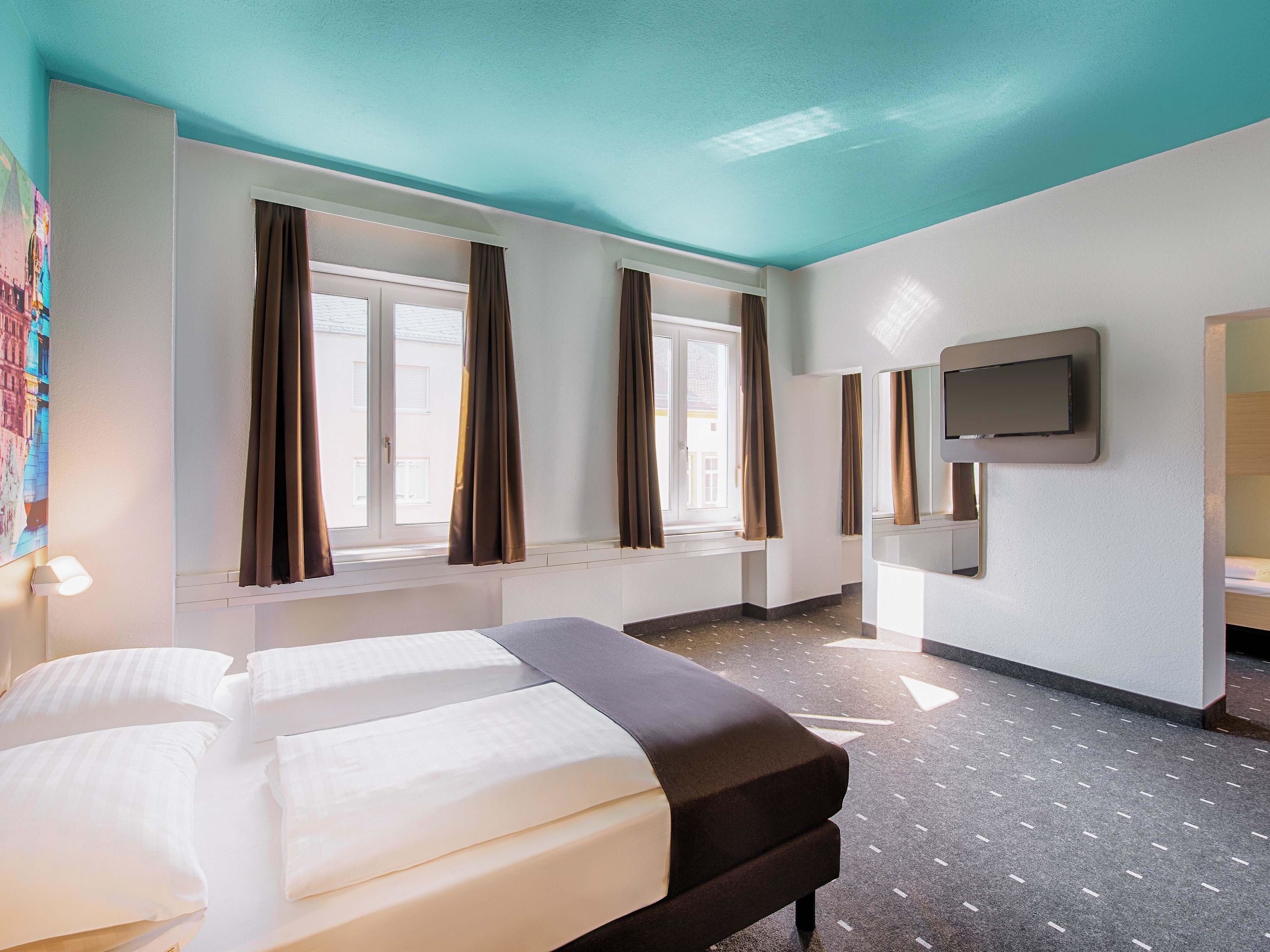 Ein Hotelzimmer mit zwei Betten, einem großen Fenster mit Vorhängen, einem an der Wand befestigten Flachbildfernseher und einem gemusterten Boden.