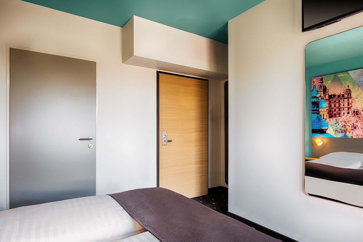 A modern hotel room with a bed, a wooden door, and a colorful cityscape mural on the wall.