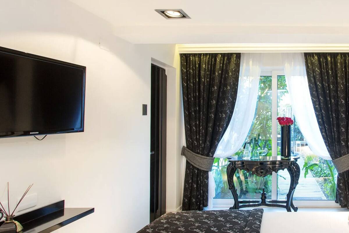 A modern hotel room with a bed, a TV, a small table with a vase, and a view of greenery outside.