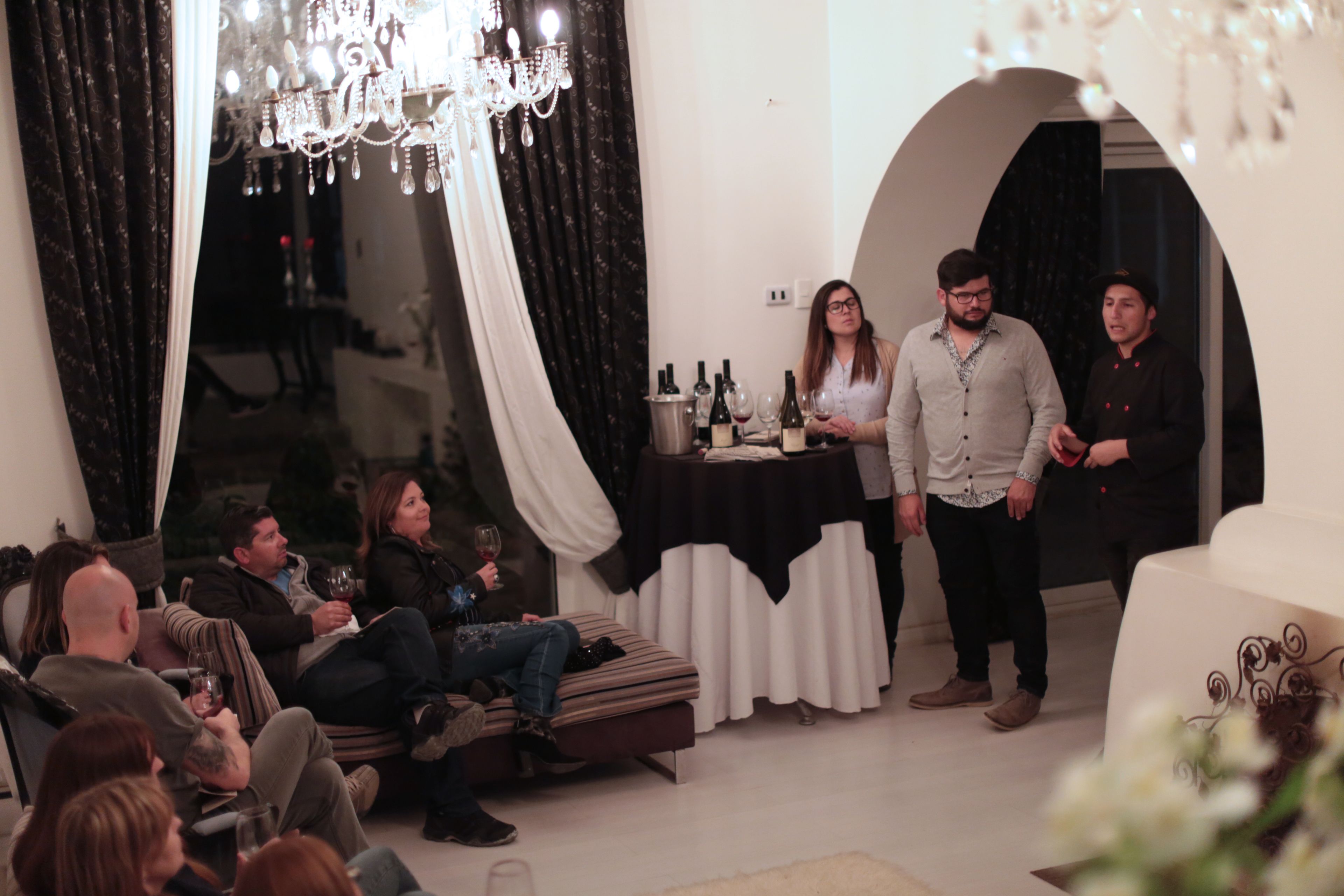 Una habitación con candelabro, una mesa con botellas y gente sentada y de pie.