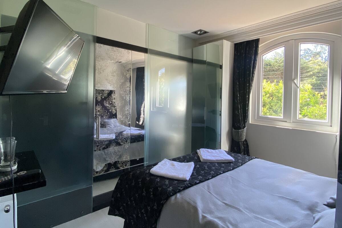A modern hotel room with a bed, a desk, a mirror, and a window with a view of greenery.