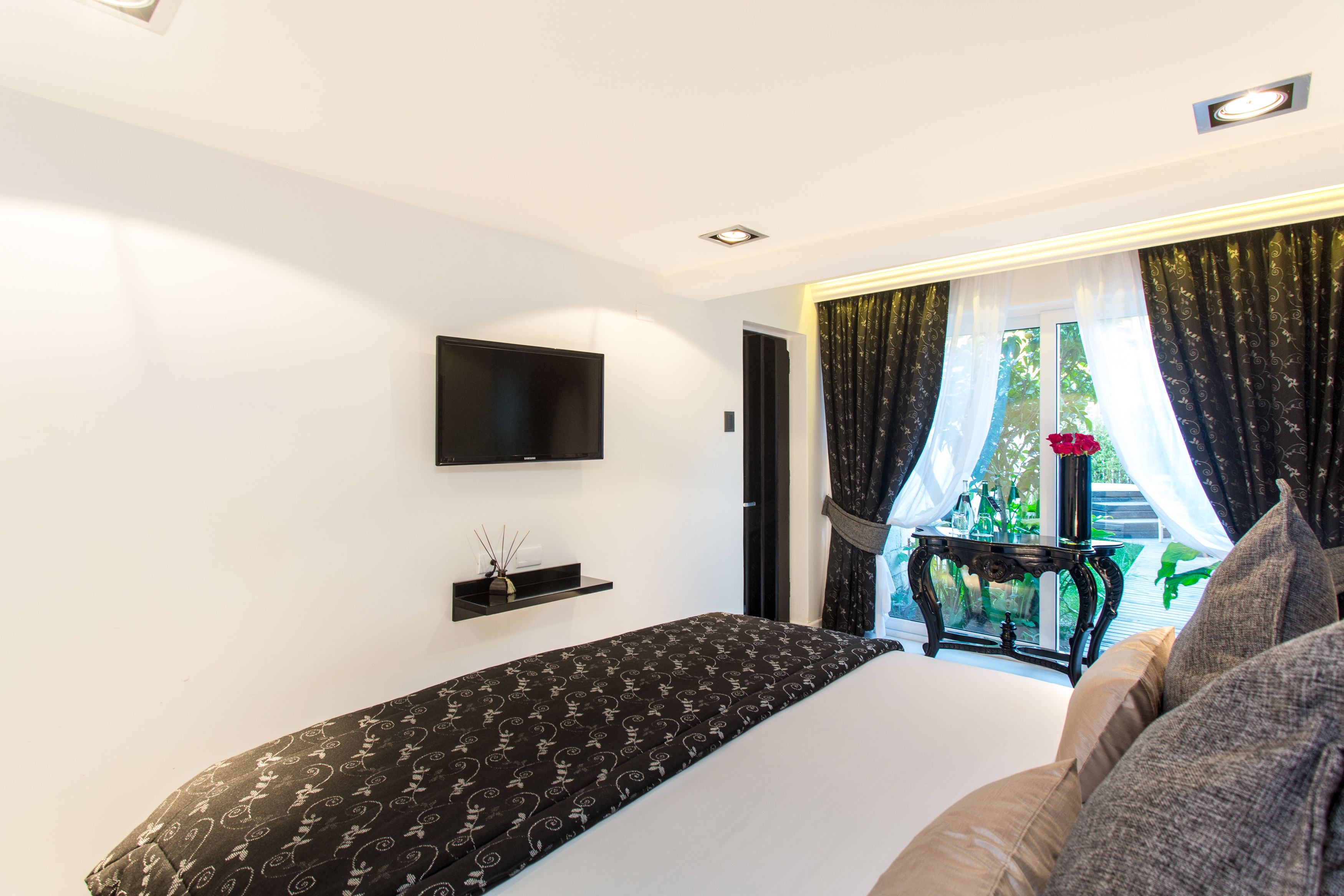 Un dormitorio moderno con una cama grande, un televisor montado, una mesita con un florero y vistas al exterior a través de cortinas translúcidas.