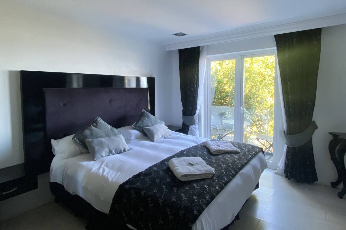 A large bed with a dark headboard, white bedding, and a patterned bedspread. There is a window with curtains and a view of greenery outside.