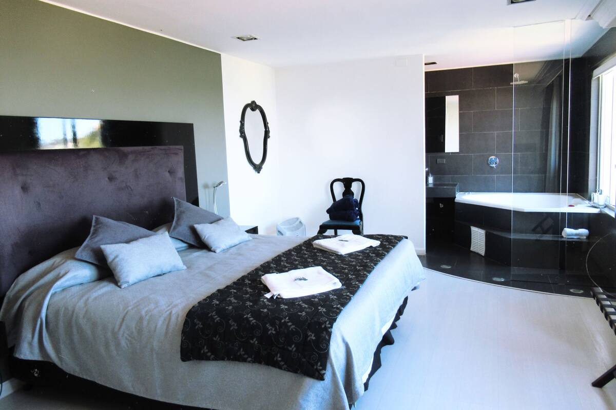 A modern bedroom with a large bed, a chair, a bathroom with a bathtub, and a shower.