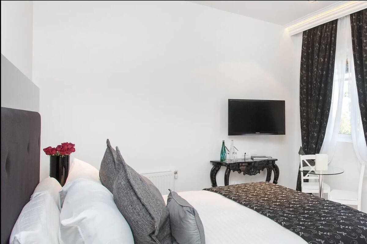 A bedroom with a bed, a TV, a small table with a vase of flowers, a chair, and a view of the outside through a window.