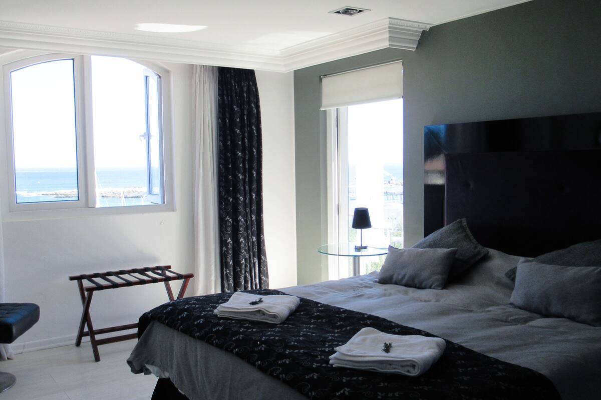 A modern hotel room with a large bed, a bench, a small table, and a view of the ocean.