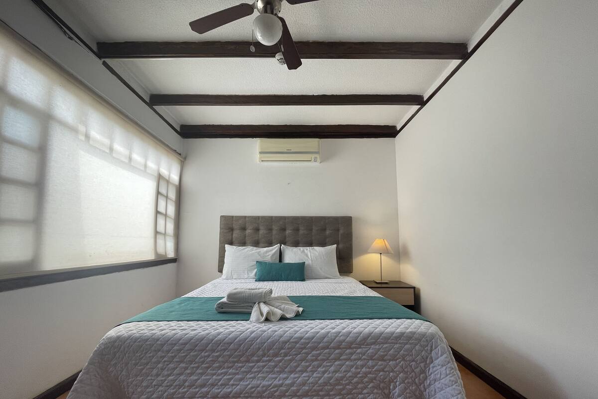 A hotel room with a bed, a ceiling fan, a nightstand with a lamp, and a window with blinds.