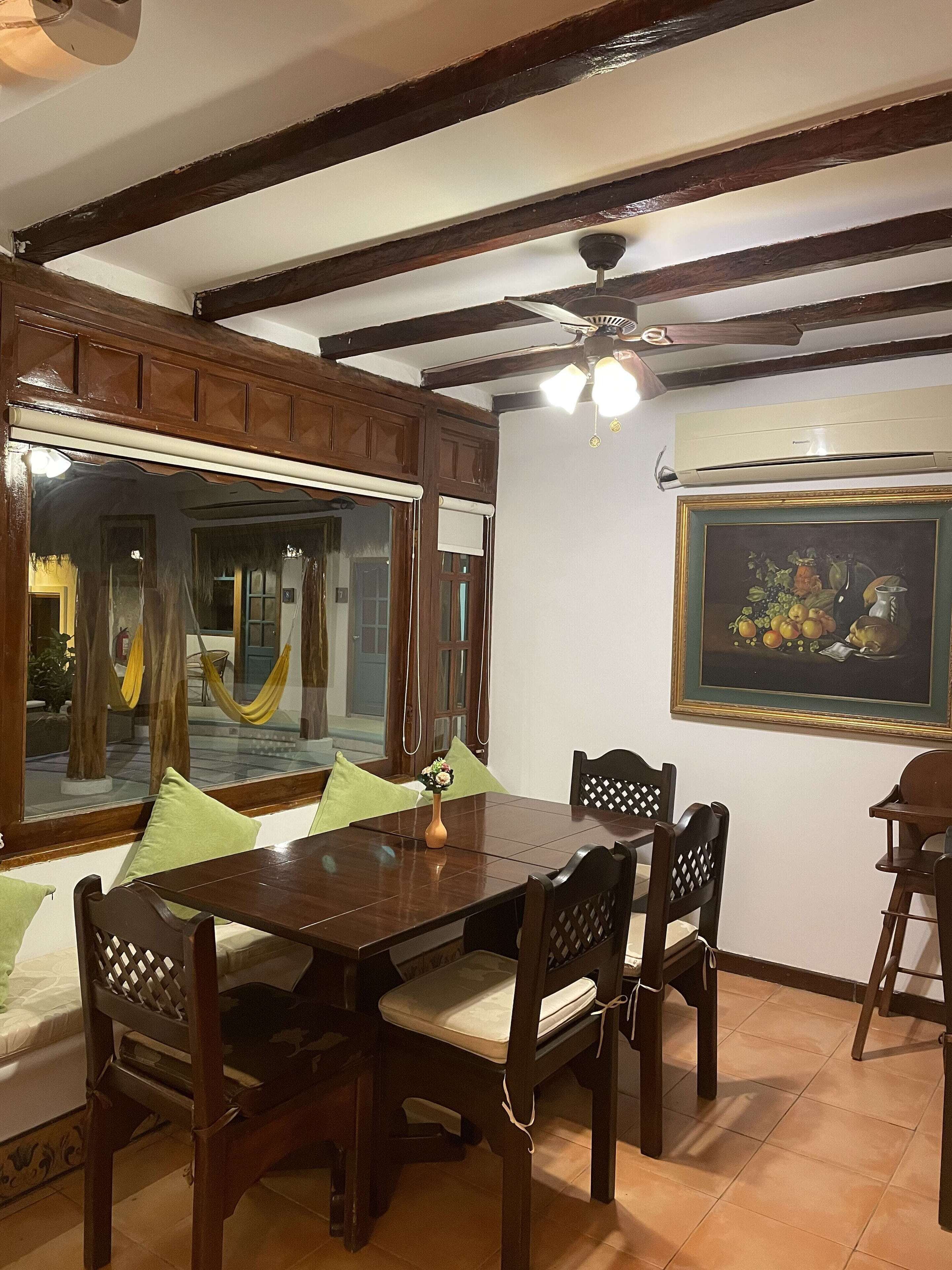 A dining area with a wooden table and chairs, a large window with a view of the outside, and a ceiling fan.