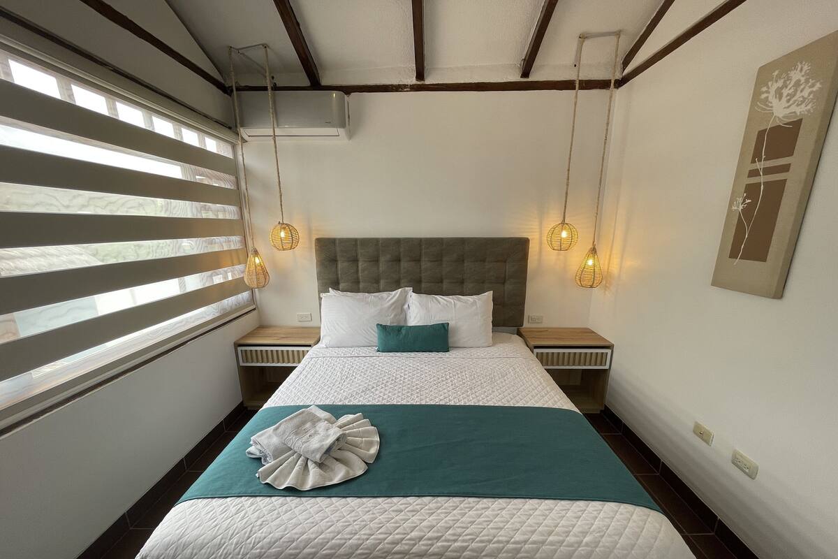 A neatly arranged hotel room with a large bed, two bedside tables, a window with blinds, and a wall-mounted air conditioner.