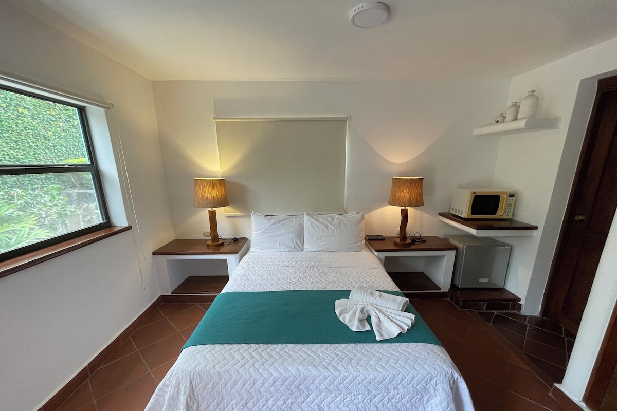 A hotel room with a bed, two bedside lamps, a microwave, and a small shelf with decorative items.