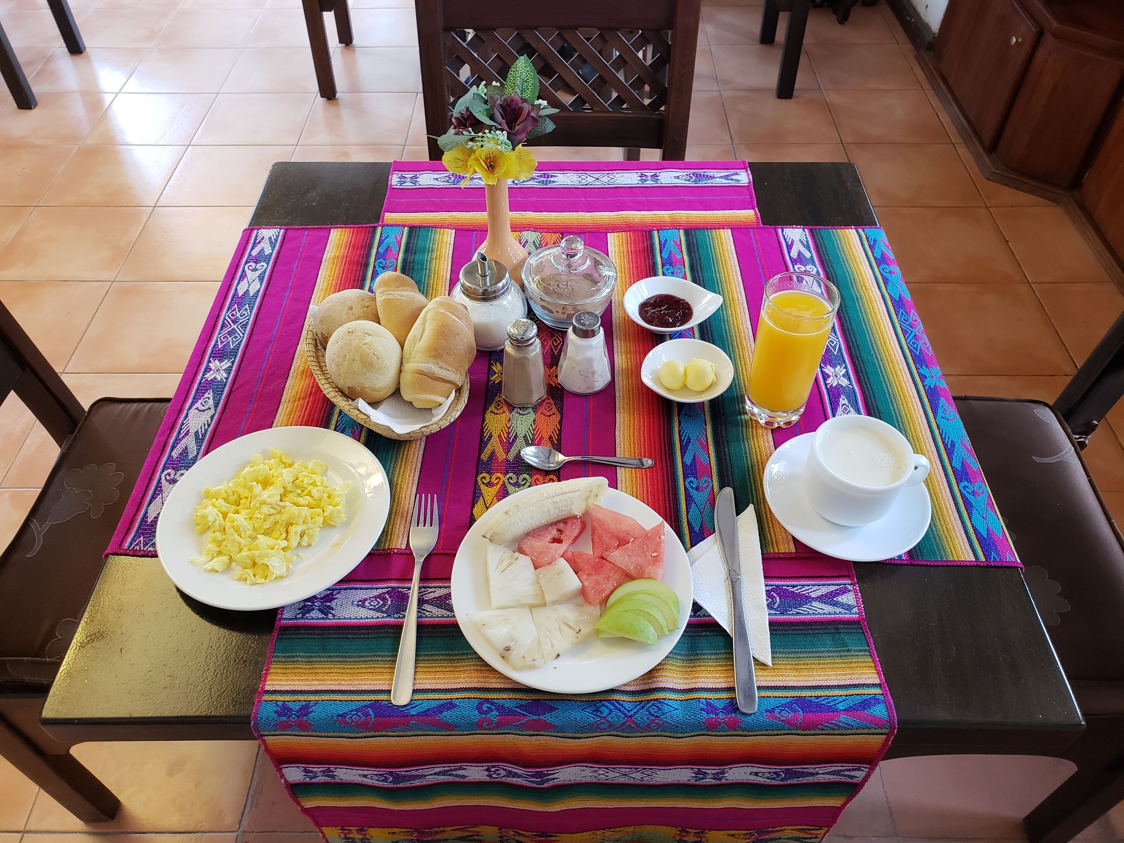 Una mesa de desayuno con platos de huevos revueltos, un plátano y una rebanada de sandía, junto con un vaso de jugo de naranja y una taza de café.