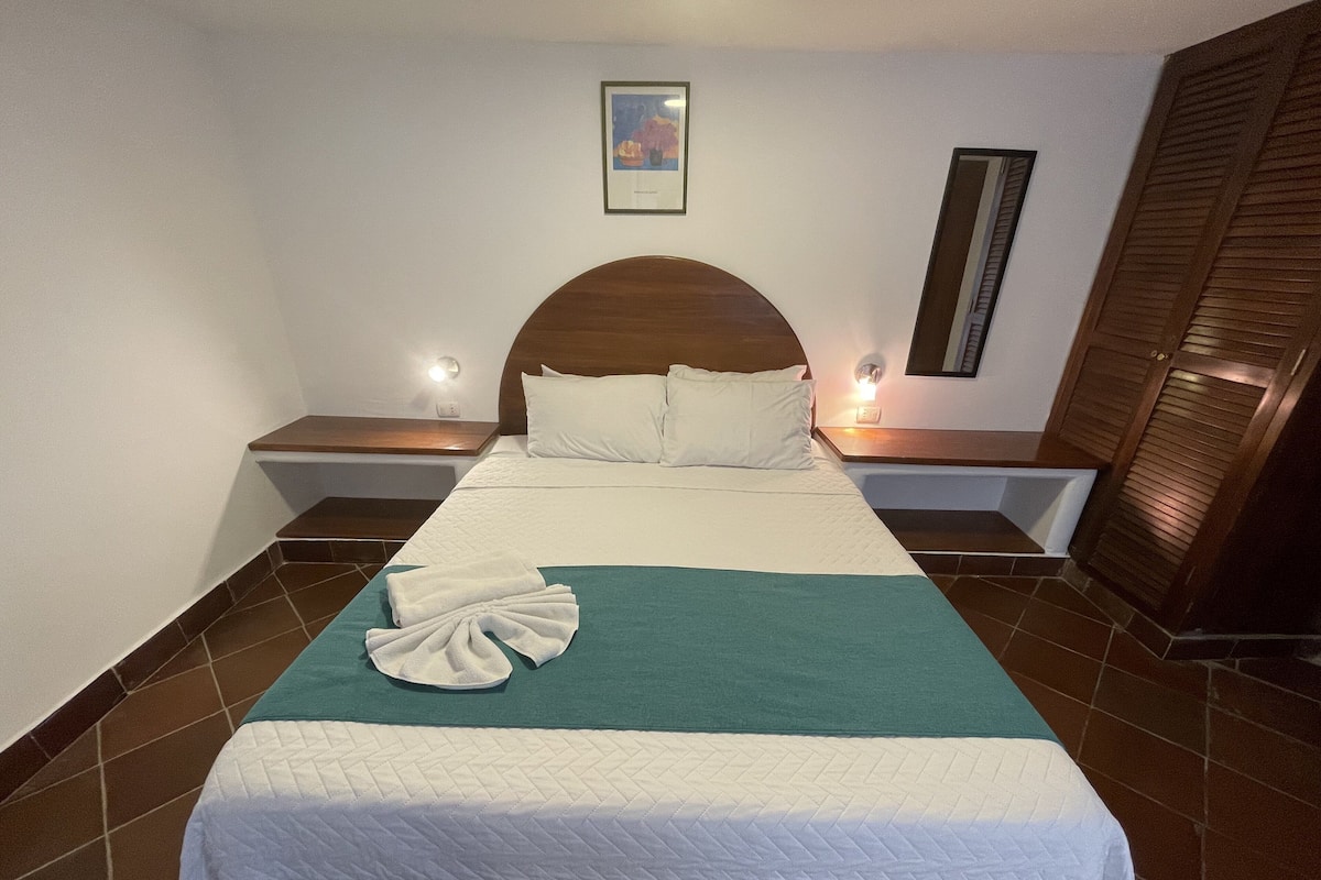 A neatly made bed with a green and white bedspread, white pillows, and a folded towel on the bed. There are two bedside tables with lamps, a wooden headboard, and a framed picture on the wall.