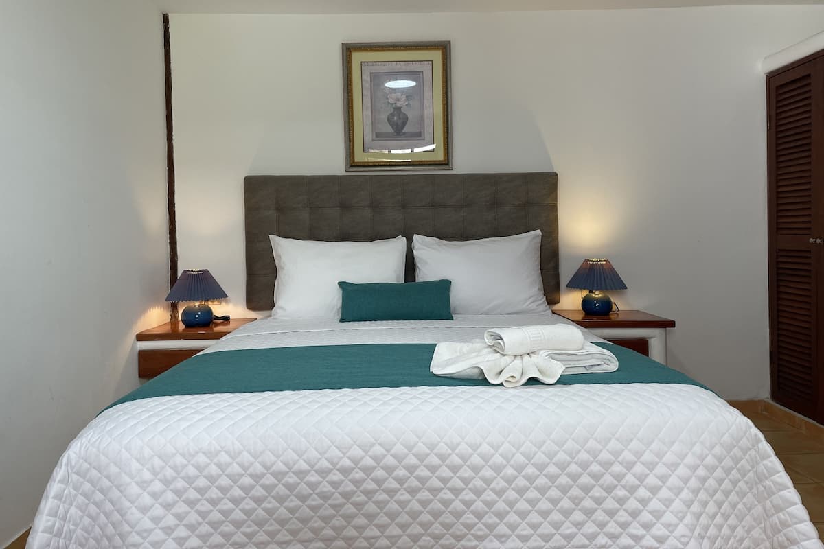 A neatly made bed with a quilted white duvet, two pillows, and a teal throw blanket. There are two bedside lamps with blue shades and a framed picture above the bed.