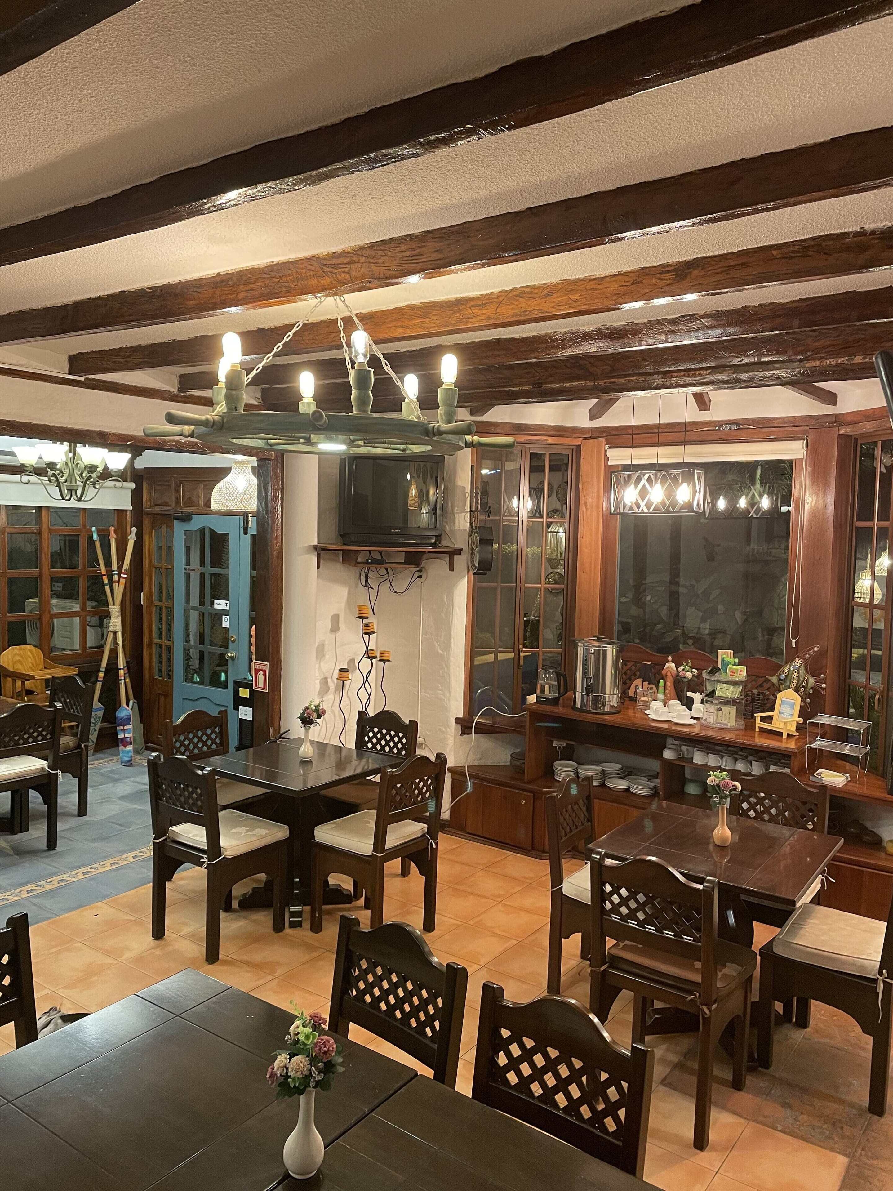 A dining area with wooden tables and chairs, a television mounted on the wall, and a chandelier above.