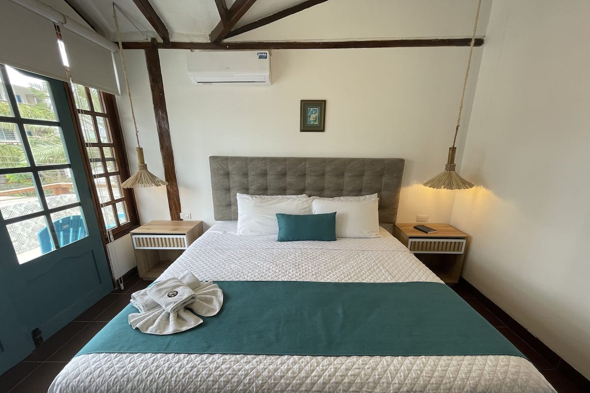 A neatly made bed with a quilted white duvet, a teal throw blanket, and a folded towel on the bed. There is a wooden nightstand with a lamp on it, and a window with a view of greenery outside.