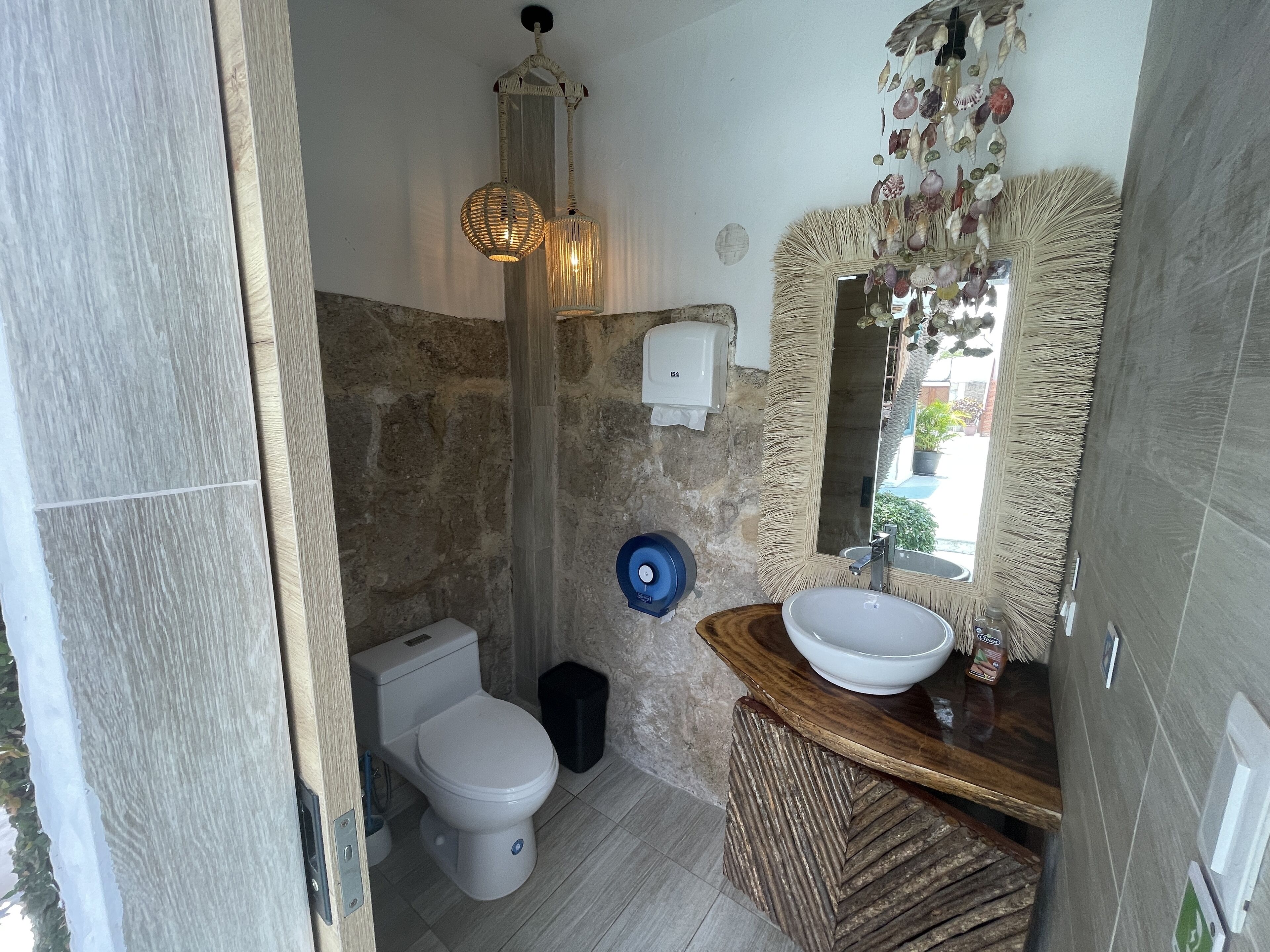 Un baño con pared de piedra, inodoro blanco, tocador de madera con lavabo y espejo con marco similar a la paja, y una lámpara colgante.