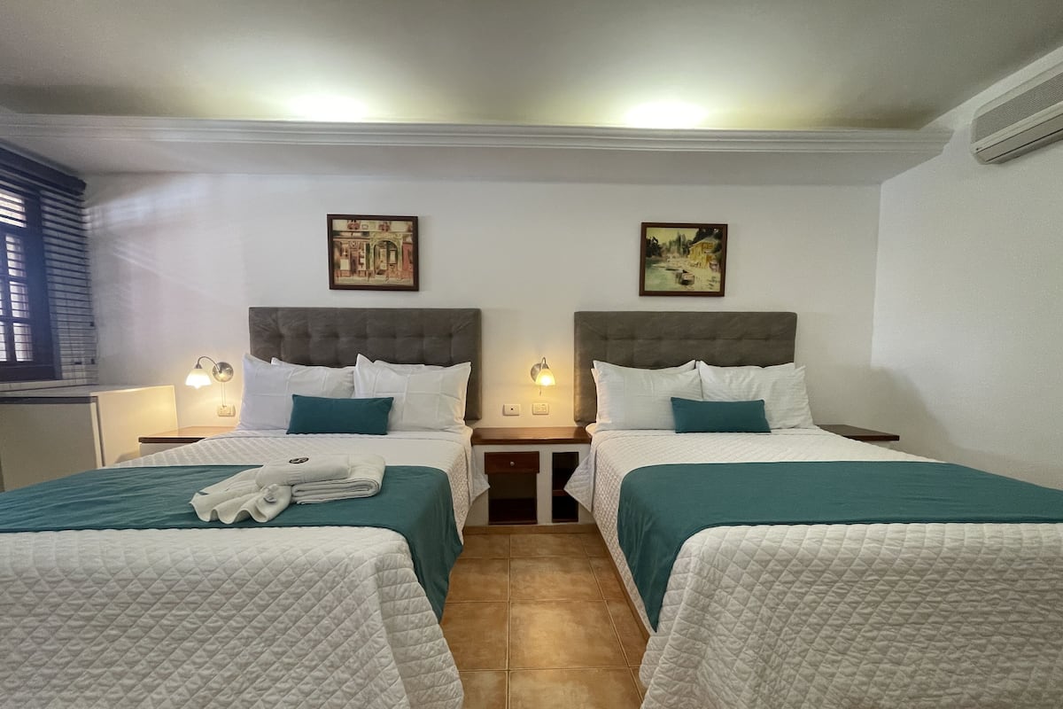 A hotel room with two beds, each with a headboard, white bedding, teal throw pillows, and a folded towel on the bed. There are two wall-mounted lamps on either side of the beds, and a painting above each bed.