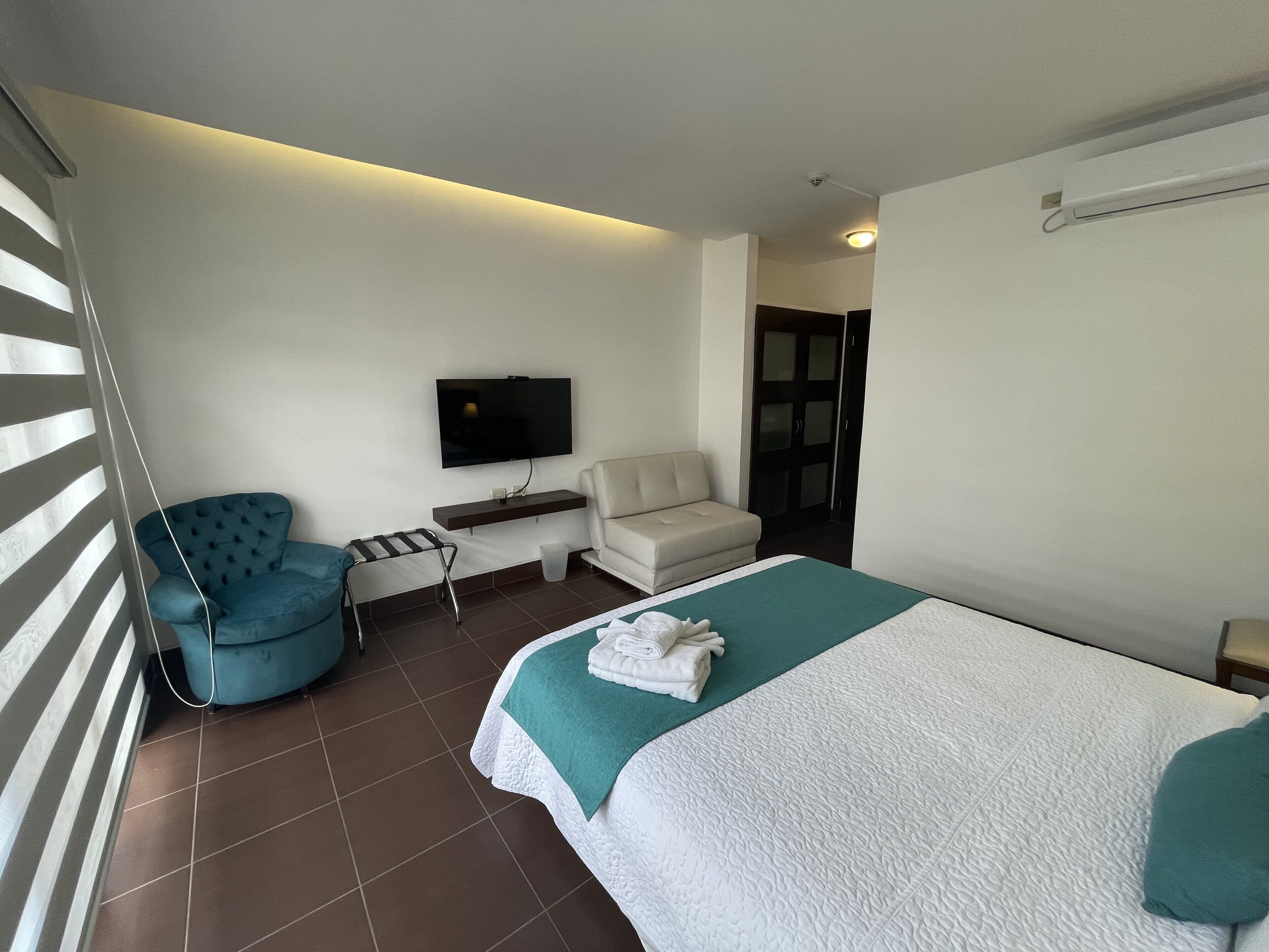 A hotel room with a bed, a blue armchair, a white sofa, a TV mounted on the wall, and a door leading to another room.