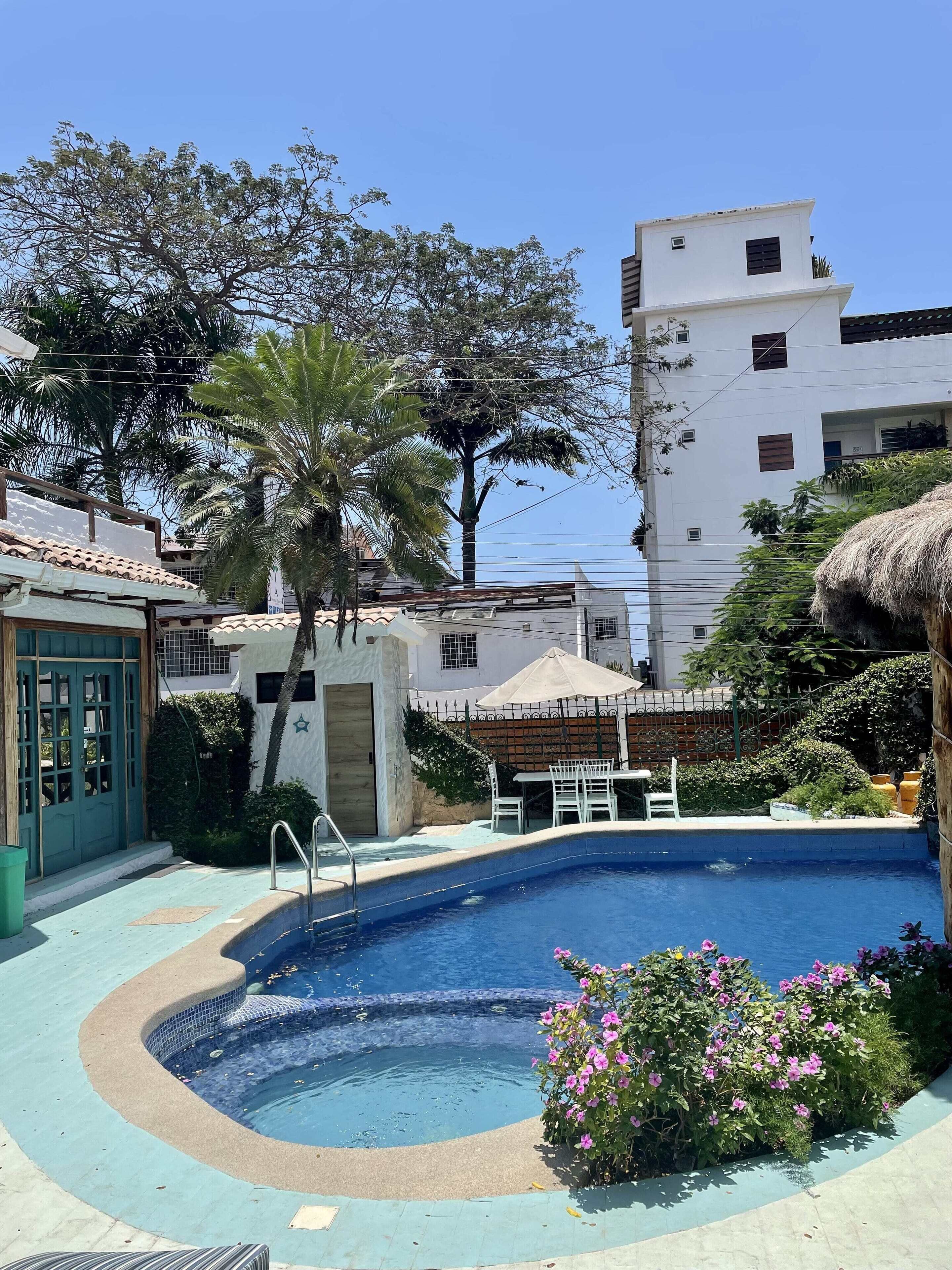 Una piscina con interior azul, rodeada por un deck azul claro. Se observan palmeras y un edificio con fachada blanca al fondo.