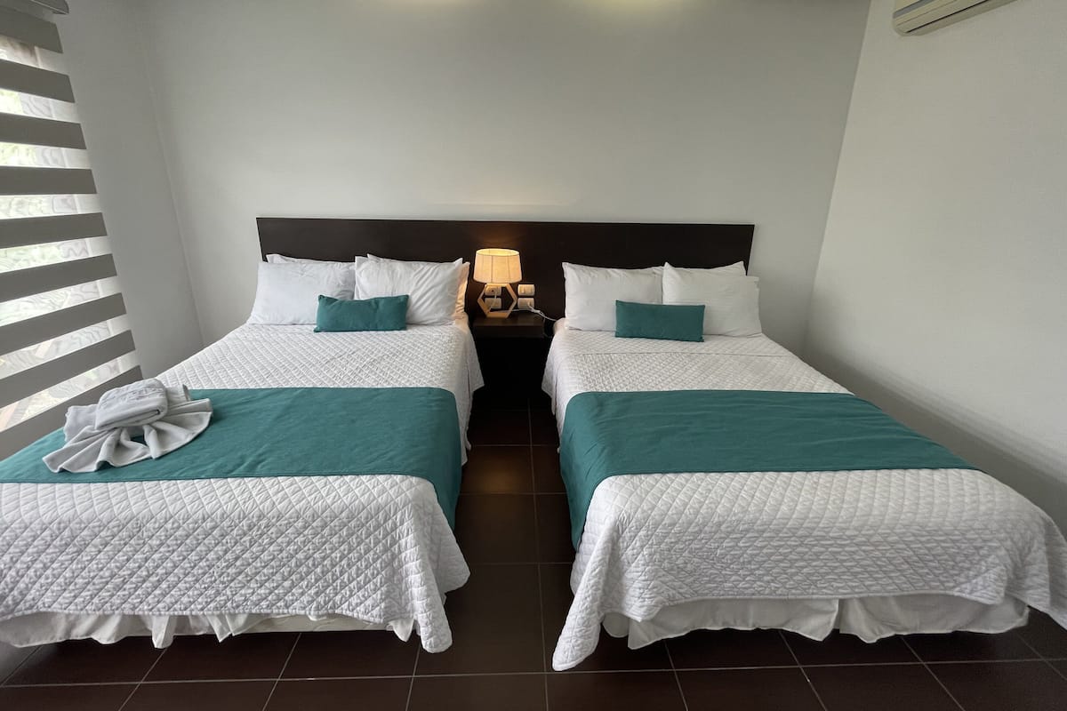 A hotel room with two beds, each with a white quilted bedspread and teal pillows, a wooden nightstand with a lamp, and a window with blinds.